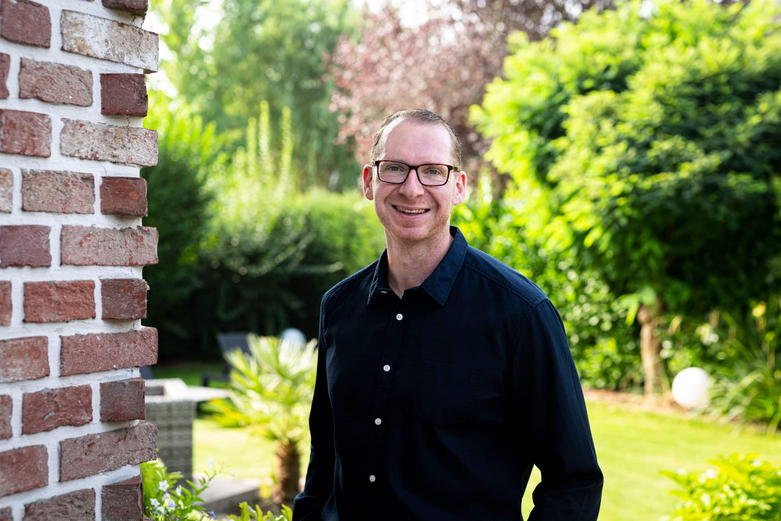 Porträt von Andreas Burdack, dem Inhaber der Immobilien, der im Garten neben einer Backsteinmauer steht.