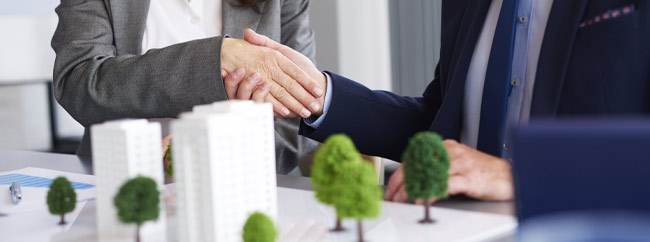 Beratung Zwei Geschäftsleute schütteln sich die Hände über einem Tisch mit Modellbäumen.
