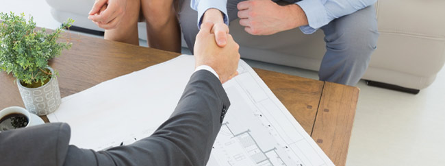 Immobilien_Zufriedenheit Zwei Geschäftsleute schütteln sich die Hände über einem Tisch mit Bauplänen.