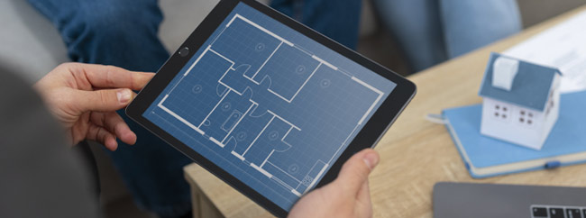 Presentation Person hält ein Tablet mit einem digitalen Grundriss eines Gebäudes, daneben ein Modellhaus auf einem Tisch.
